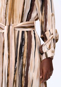 Vloeiende jurk met gestreepte patronen in beige, bruin en zwart. Beschikt over een ceintuur in de taille, knopen op de manchetten en zachte, textuurstof.