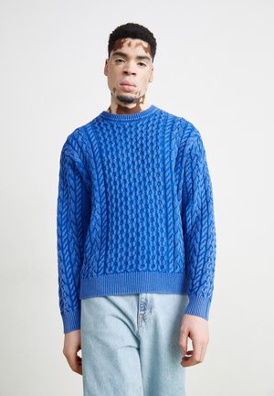 Blue knitted sweater featuring a textured cable pattern, round neckline, and ribbed cuffs and hem. Worn over light blue denim jeans.