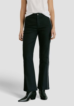 Vrouw draagt zwarte hooggetailleerde broek met zijstreep, gecombineerd met een witte mouwloze top en zwarte enkellaarsjes met puntige neus.