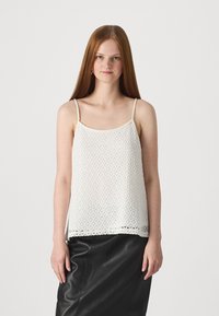 Hvit heklede tank-top med tynne stropper, med et åpent vevemønster og en bølget blonderkant, kombinert med et svart skinnskjørt.