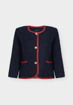 Femme aux cheveux blonds courts portant une veste bleu marine avec un liseré rouge, une chemise blanche, un jean foncé, des talons beiges et tenant un petit sac à main rouge.