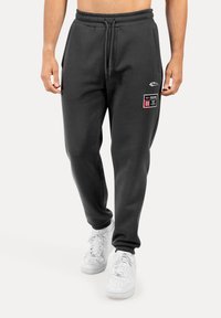 Šedé tepláky vyrobené z měkké látky, mají stahovací pas a dvě kapsy. Na levé noze je viditelná grafická nášivka. Bílá obuv doplňuje celý vzhled.