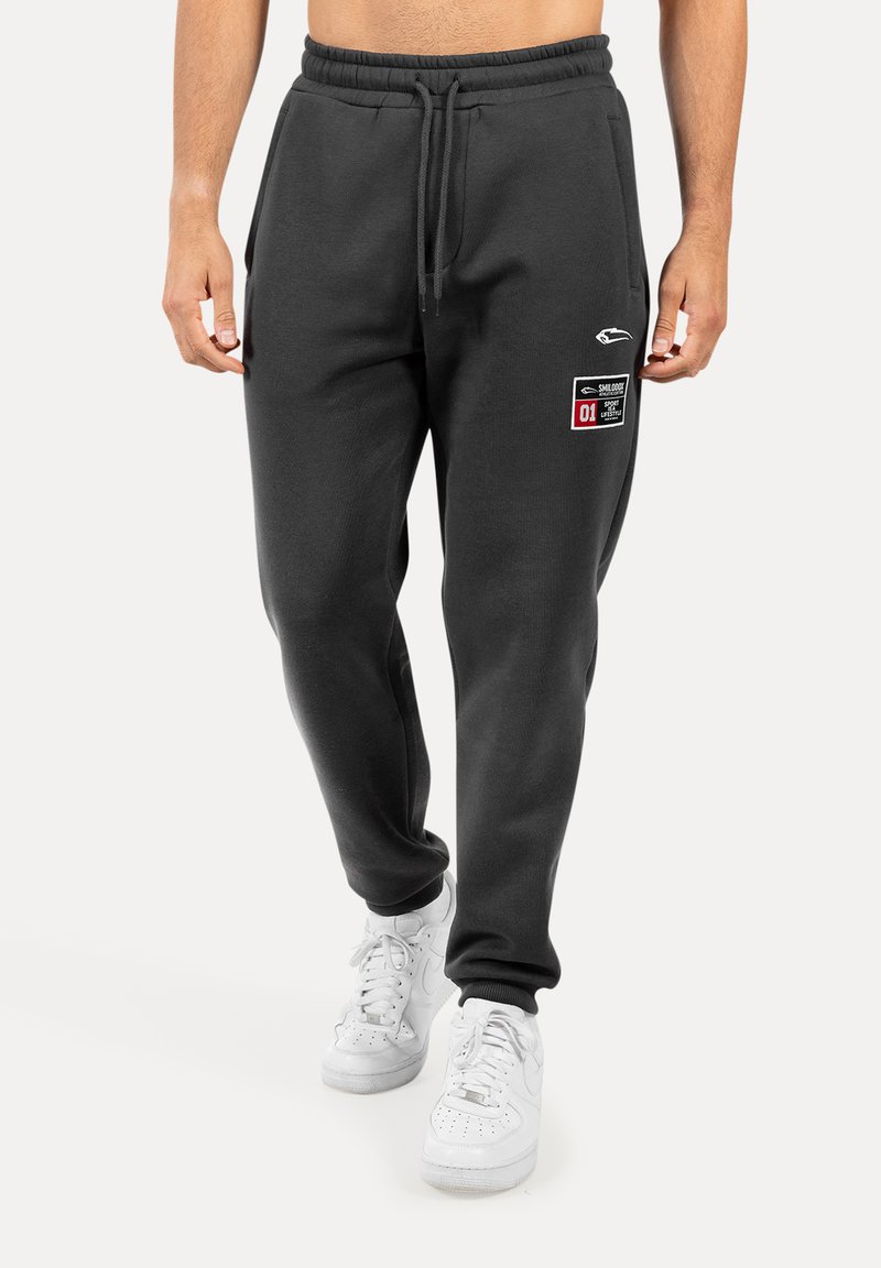 Šedé tepláky vyrobené z měkké látky, mají stahovací pas a dvě kapsy. Na levé noze je viditelná grafická nášivka. Bílá obuv doplňuje celý vzhled.