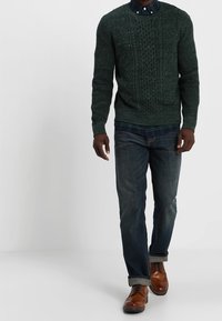 Dunkelgrüner Kabelstrickpullover mit Rundhalsausschnitt über einem karierten Hemd, kombiniert mit dunklen Jeans und braunen Lederschuhen.