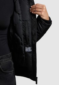 Geacă neagră pufată cu textură matlasată, buzunare cu fermoar și o etichetă interioară cu inscripția „Khujo Original Changing Threads”.