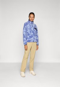 Blauer Fleece-Pullover mit abstraktem Muster, halber Knopfleiste und schwarzem Brusttasche. Kombiniert mit Khaki-Hosen und beigen Sneakern.