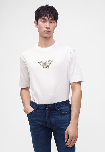 Νέος άνδρας με λευκό t-shirt με λογότυπο αετού και σκούρα μπλε τζιν, στέκεται με το ένα χέρι στην τσέπη του μπροστά από ένα απλό φόντο.