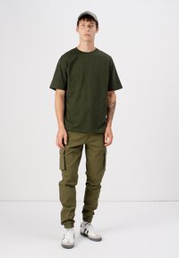 Olive green cotton t-shirt, relaxed fit, paired with olive cargo pants featuring multiple pockets. Worn with gray cap and white sneakers.