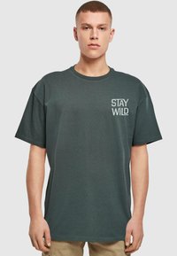 Tmavě zelené tričko s krátkým rukávem vyrobené z bavlny, s bílým potiskem "STAY WILD" na levé hrudi. Uvolněný střih, design s kulatým výstřihem.