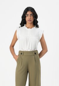 Blusa bianca con maniche a balze e dettagli in pizzo all'orlo, abbinata a pantaloni verdi oliva a vita alta con due bottoni neri.
