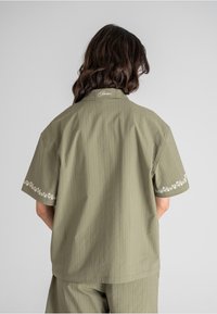 Olivenfarget kortermet skjorte med broderte blomstermotiver på ermene og kragen; teksturert stoff med vertikale pinstriper.