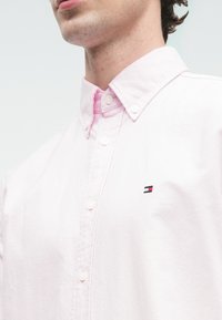 Gaiši rozā pogājama krekls ar apkaklīti. Uz krūšu daļas ir neliels zīmola logo ar sarkaniem, baltiem un ziliem akcentiem. Gluda tekstūra.