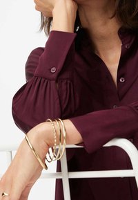 Bordeauxfarvet bluse med puffede ærmer og knapmanchetter, der har en glat tekstur. Guldarmbånd båret på håndleddet, som står i kontrast til stoffet.