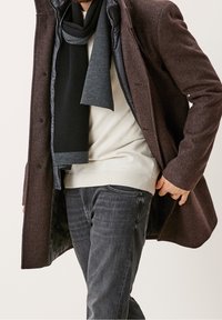 Cappotto marrone strutturato con foderato nero imbottito, abbinato a un maglione chiaro e jeans in denim scuro. Accessoriato con una sciarpa nera.