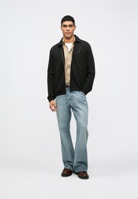 Crna jakna, beži majica, plavi široki traperice i tamnosmeđe cipele. Jakna ima kragnu i diskretne prednje džepove.