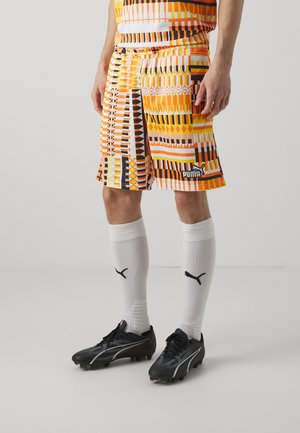 Pantaloni corti in arancione e giallo vivace con motivi geometrici, abbinati a calzini bianchi e scarpe da calcio nere con dettagli argentati.