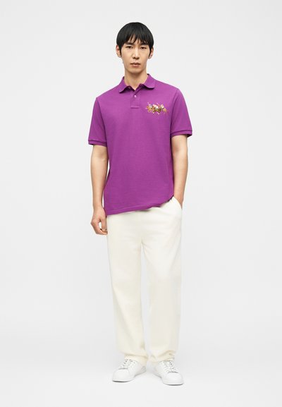 Άνδρας με μοβ πόλο πουκάμισο με κεντημένη στάμπα παίκτη πόλο, άσπρο παντελόνι και άσπρα αθλητικά παπούτσια, στέκεται με το ένα χέρι στην τσέπη.