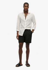 Camicia di lino a maniche lunghe bianca, pantaloni da bagno neri con logo e sandali neri a ciabatta. Il modello è in piedi di fronte.