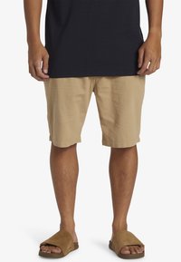 Beige bomullsshorts med uppvikta fållar, kombinerade med bruna sandaler. Modellen bär en mörk t-shirt och står framför en neutral bakgrund.