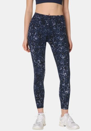 Femme portant des leggings bleu marine à motifs et taille haute ainsi que des chaussures de sport blanches, debout devant un fond clair uni.