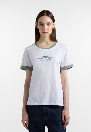 Donna con capelli castani lisci indossa una maglietta bianca con bordi verdi e la scritta "Summer Diaries" con una grafica di racchette da tennis incrociate.