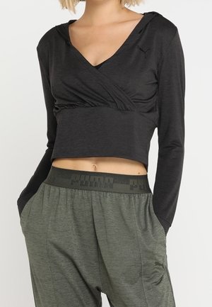 Vrouw die een zwart crop top met lange mouwen en een V-hals draagt, en een grijze Puma joggingbroek met handen in de zakken.