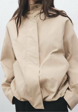 Frau trägt eine beige locker sitzende Jacke mit hohem Kragen und verdecktem Frontverschluss, die Hände in den Seitentaschen vergraben.