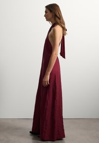 Burgunderfarbene Maxikleid mit crinkled Textur, Halterneck-Design und offener Rückseite. Mit einer gebundenen Akzentuierung am Ausschnitt und einer fließenden Silhouette.