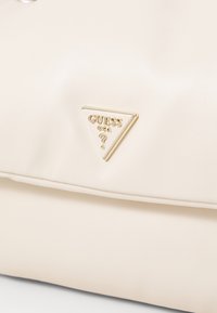 Superficie in pelle color crema con un logo triangolare Guess USA bordato d'oro, caratterizzato da un punto interrogativo al centro.