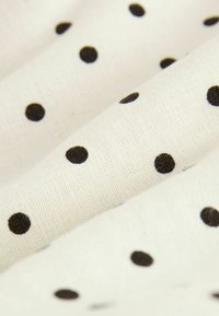 Tissu blanc à texture tissée présentant des petits pois noirs espacés de manière régulière, montrés en plis doux.