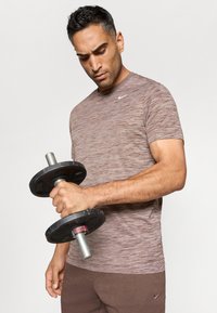 Hombre vestido con ropa deportiva marrón levantando una mancuerna negra, concentrado en su entrenamiento de brazos, contra un fondo claro y sencillo.