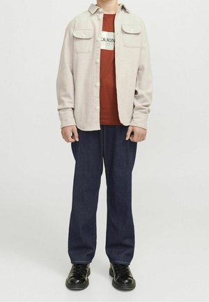 Persona con camisa beige abotonada sobre camiseta naranja quemado, pantalones rectos azul oscuro y zapatos negros con cordones, de pie frente a un fondo liso.