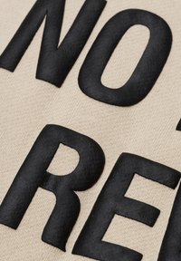 Platneno platno u bež boji s velikim, podebljanim crnim slovima. Slova imaju glatku, teksturiranu površinu koja se kontrastira s pozadinom.