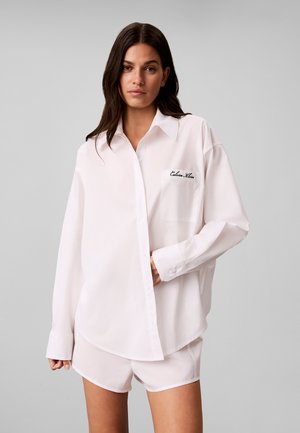 Mujer con cabello largo y oscuro, vestida con una camisa blanca suelta abotonada y shorts a juego sobre un fondo liso gris claro.