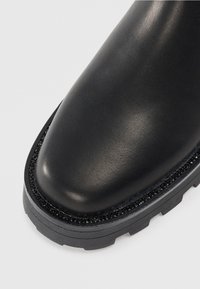 Detailní záběr na černou koženou botu s kulatým čtvercovým špičkou, ozdobnými černými hroty kolem podrážky a silnou gumovou profilovanou podrážkou.