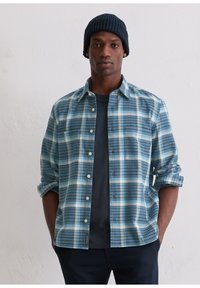 Koszula w kratę w odcieniach niebieskiego i białego, z długim rękawem, zapinana na guziki, z kieszenią na piersi, noszona na ciemnogranatowej koszulce.