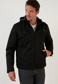 Svart zip-up jacka med avtagbar huva, texturerade axelpaneler och framfickor. Tillverkad av ett glatt, något glänsande tyg.