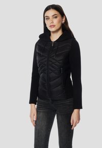 Črna puhovka z zadrgo spredaj, diamantnim vzorcem in mehko kapuco. Gumbi in žepi imajo kontrastno gladko tkanino.