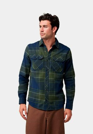 Camisa de franela a cuadros en verde oscuro y azul marino, con dos bolsillos en el pecho, cierre de botones en la parte delantera y mangas largas con un cuello estructurado.