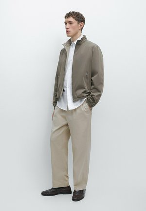Giovane uomo con pantaloni beige, camicia bianca con bottoni, giacca taupe con cerniera e mocassini marrone scuro, in piedi contro uno sfondo chiaro e semplice.