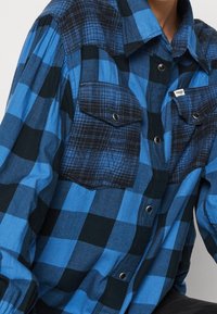 Blue and black checkered flannel shirt with two chest pockets, snap buttons, and a mixed pattern design. Soft fabric with a textured finish.