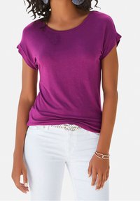 Fialový krátký rukáv tričko s ohrnutými manžetami, hladká látka, kulatý výstřih, nošeno přes bílé džíny s pleteným páskem a náramkovým detailem.