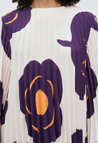 Žena nosi bielu plisovanú blúzku s veľkými fialovými a oranžovými abstraktnými kvetinovými vzormi viditeľnými od ramien po stred trupu.