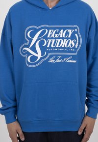 Man in een blauwe hoodie met op de borst het witte logo "Legacy Studios Automobile, Inc. Live Fast & Curious" en met ontspannen handen aan de zijkanten.