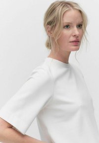 Valkoinen lyhythihainen paita, jossa on korkea kaulus, sileä pinta ja rakenteellinen istuvuus, esillä yksinkertaisella vaalealla taustalla.