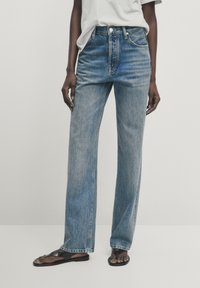 Ljusblå denimjeans med rak bendesign, hög midja, framträdande sömmar och femfickors stil. I sällskap med svarta sandaler.