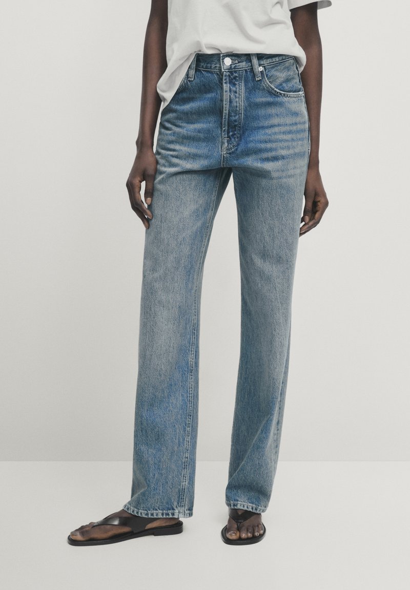 Ljusblå denimjeans med rak bendesign, hög midja, framträdande sömmar och femfickors stil. I sällskap med svarta sandaler.