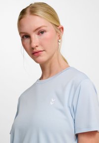 T-shirt bleu clair à manches courtes en tissu doux, avec un petit emblème blanc sur la poitrine. Encolure ronde classique et coupe décontractée.