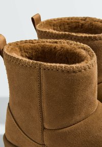 Gros plan de bottes d’hiver en daim marron avec une doublure intérieure douce et duveteuse visible et des bords supérieurs cousus sur un fond uni.