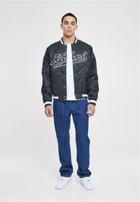 Black quilted bomber jacket with white accents and logo over a white shirt, paired with blue denim jeans and white sneakers.